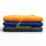 Three folded microfiber cleaning towels stacked vertically on a white background, in orange, blue, and dark gray.