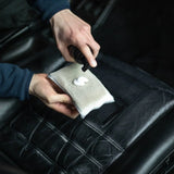 A close-up of a person's hands holding a gray microfiber cleaning cloth with a dollop of white foam, spraying a black leather car seat.