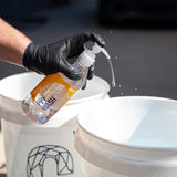 Person wearing black gloves holding a bottle labeled 'PPT Wash' over two white buckets.