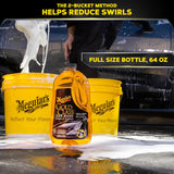 Meguiar's Gold Class Car Wash bottle displayed in front of two yellow Meguiar's buckets on a wet car, with foam being poured onto the surface. Text overlays read 'THE 2-BUCKET METHOD HELPS REDUCE SWIRLS' and 'FULL SIZE BOTTLE, 64 OZ'.